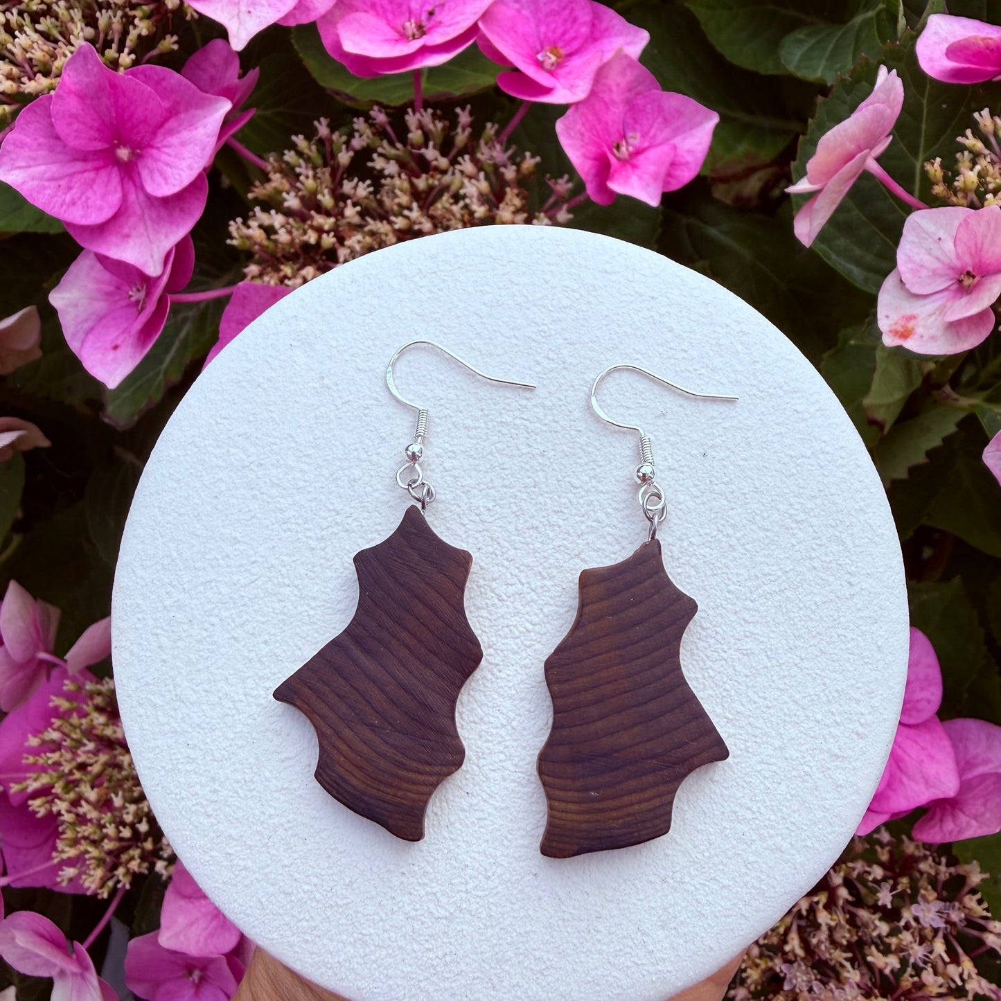 A pair of dangling earrings with dark brown wood-grain shaped pendants and silver fishhook wires, displayed on a round white stand surrounded by pink flowers and green leaves.