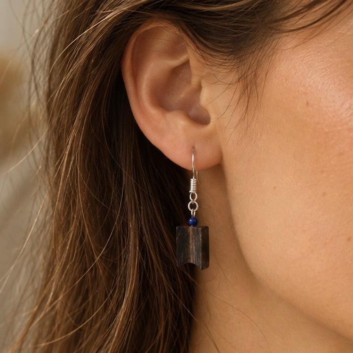 A close-up of a person's ear wearing a dangling earring. The earring has a silver fishhook wire, a small dark blue bead, and a dark rectangular wooden charm.