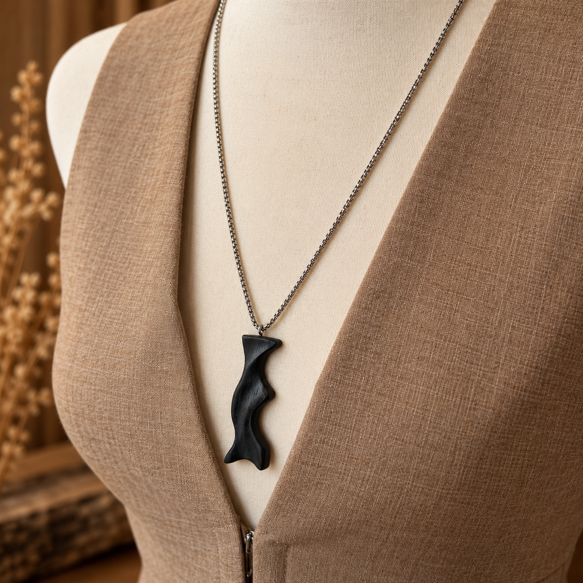 A close-up of a necklace displayed on a beige mannequin torso wearing a brown linen vest. The necklace features a textured, organic-shaped black pendant on a silver-toned chain.