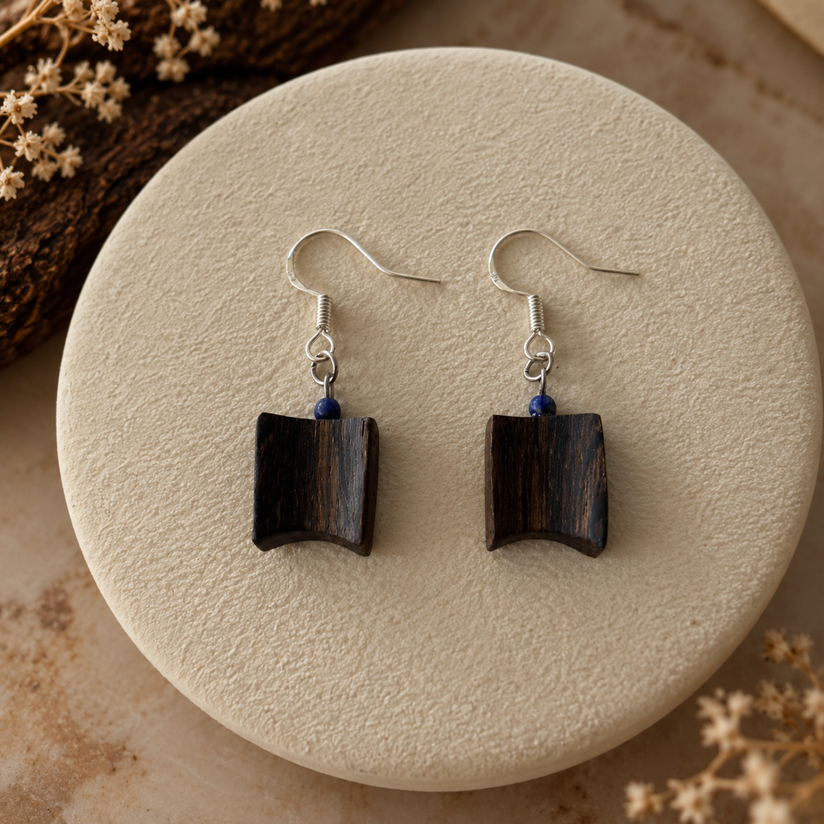 A pair of dark wood geometric dangle earrings with silver fishhook wires and small blue bead accents, displayed on a round beige textured stone plate with dried floral props.