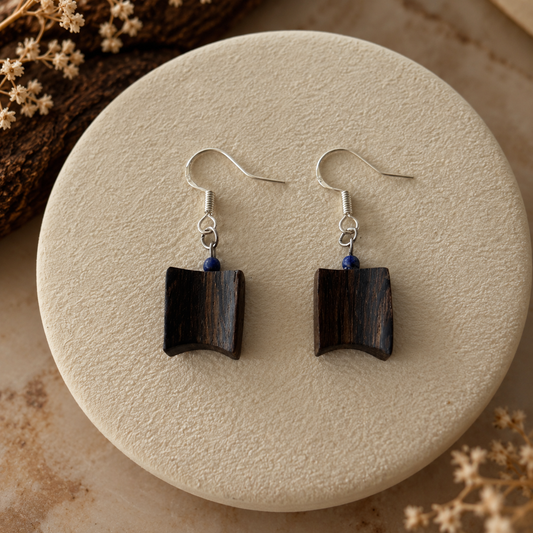 A pair of dark wood geometric dangle earrings with silver fishhook wires and small blue bead accents, displayed on a round beige textured stone plate with dried floral props.