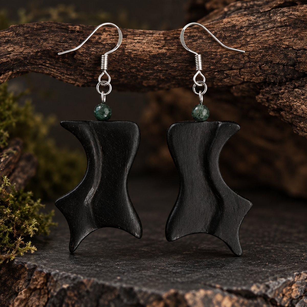 A pair of handmade black wooden dangle earrings with silver fishhook wires and small green beads, displayed on a dark textured surface with a rustic wood branch background.