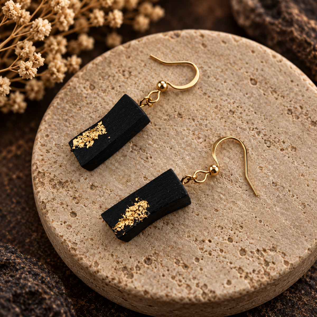 A pair of black rectangular drop earrings with gold foil leaf accents and gold fishhook ear wires, displayed on a round beige stone plate with dried flowers in the background.