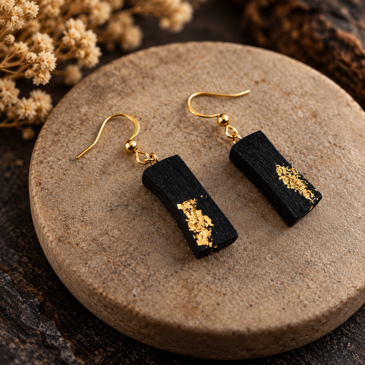A pair of black rectangular drop earrings with textured gold leaf accents and gold fishhook ear wires, displayed on a round stone coaster with dried flowers in the background.