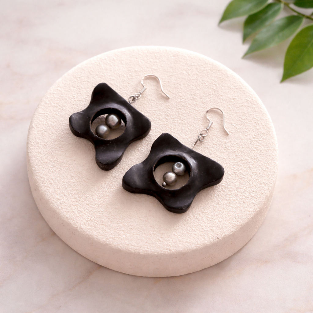 Black floral earrings on a round white stone surface with a leafy green plant in the background.