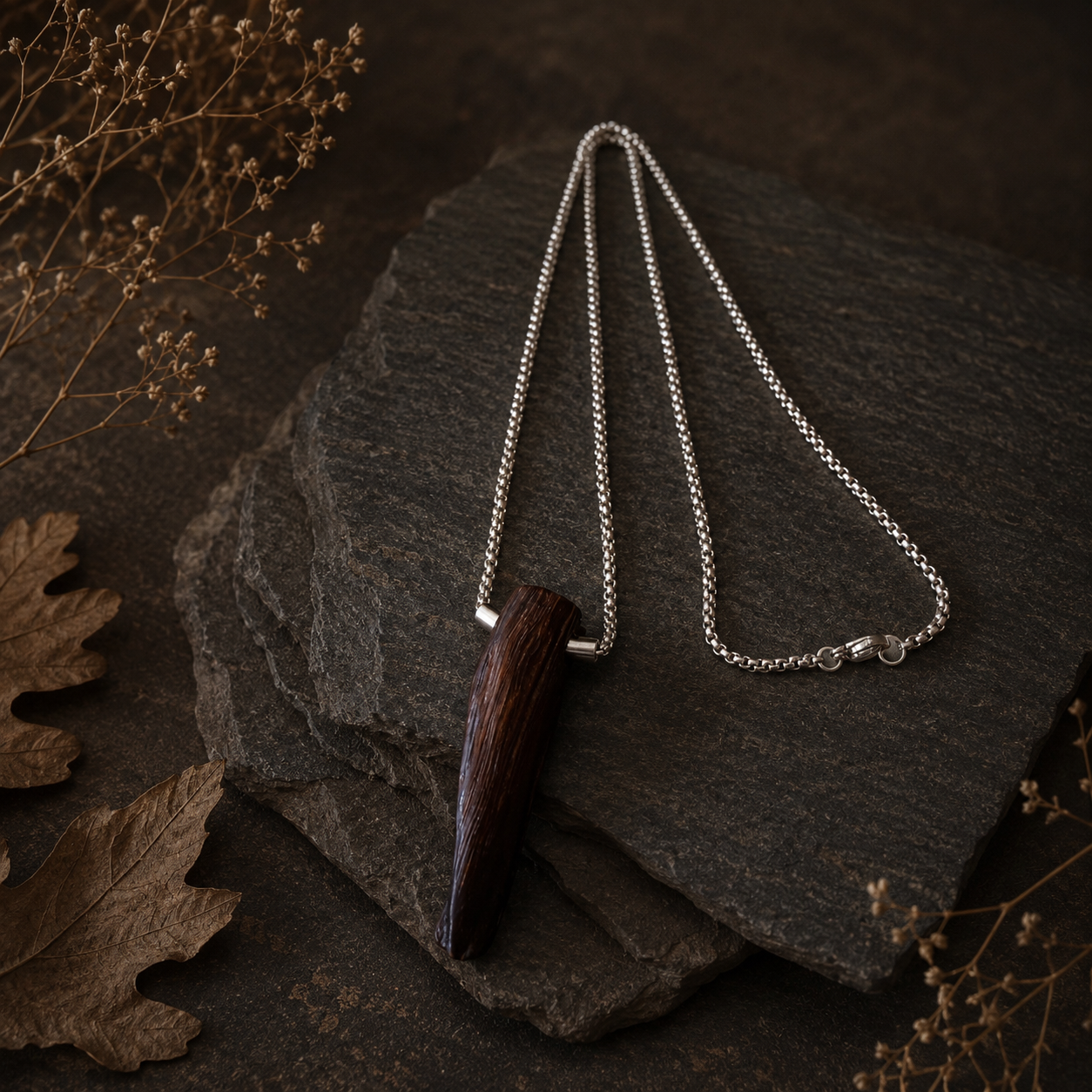 Necklace with a wooden pendant on a dark stone surface with dried leaves and flowers.