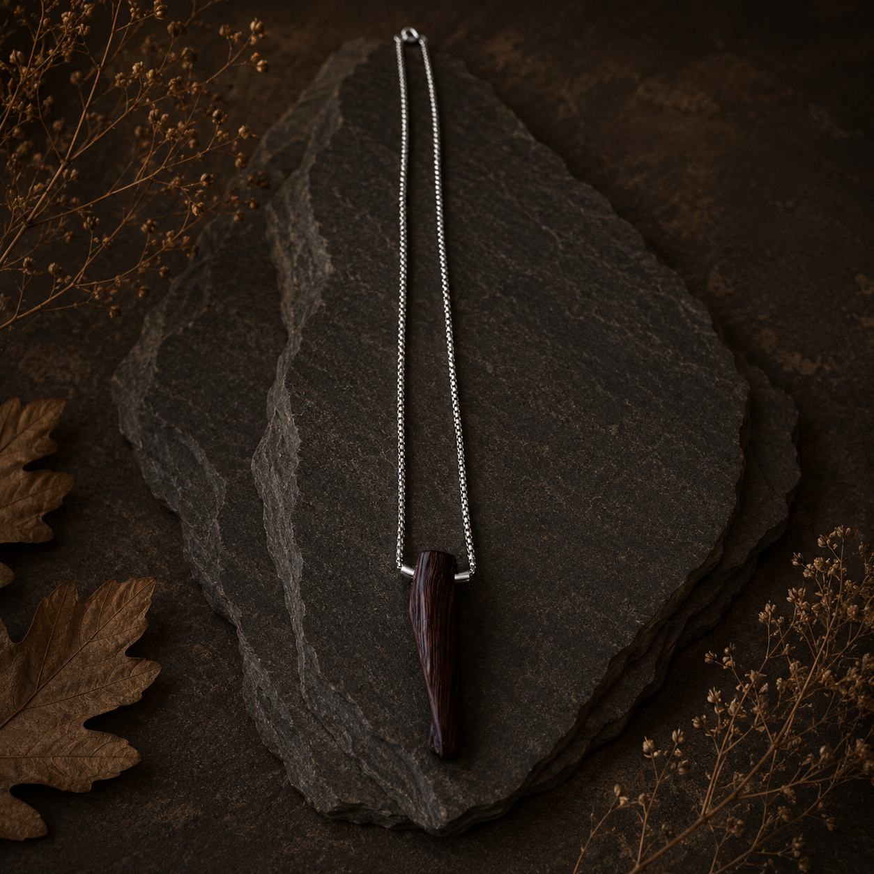 Necklace with a dark pendant on a dark stone surface with dried leaves and flowers.