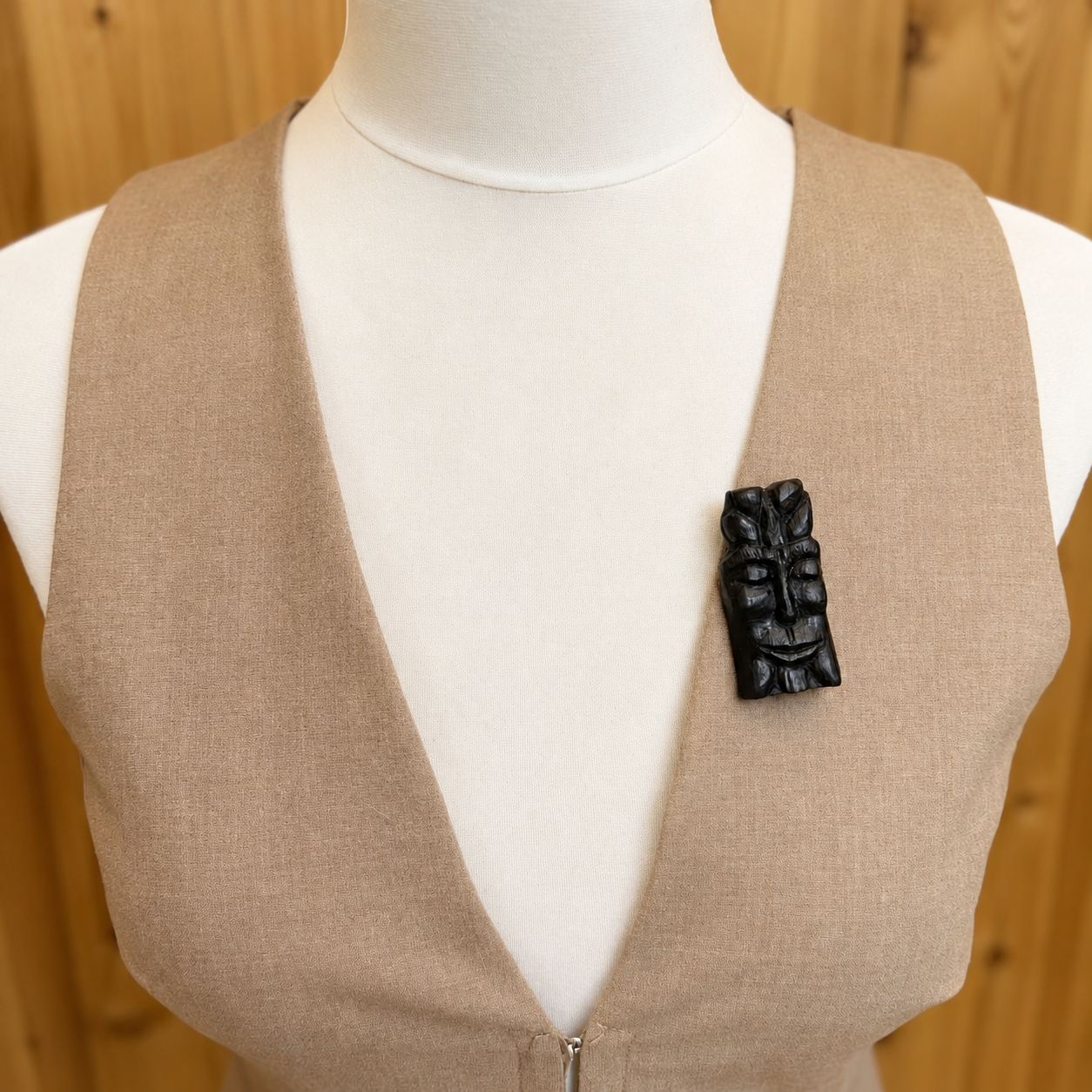 Mannequin wearing a beige vest with a black decorative brooch against a wooden background