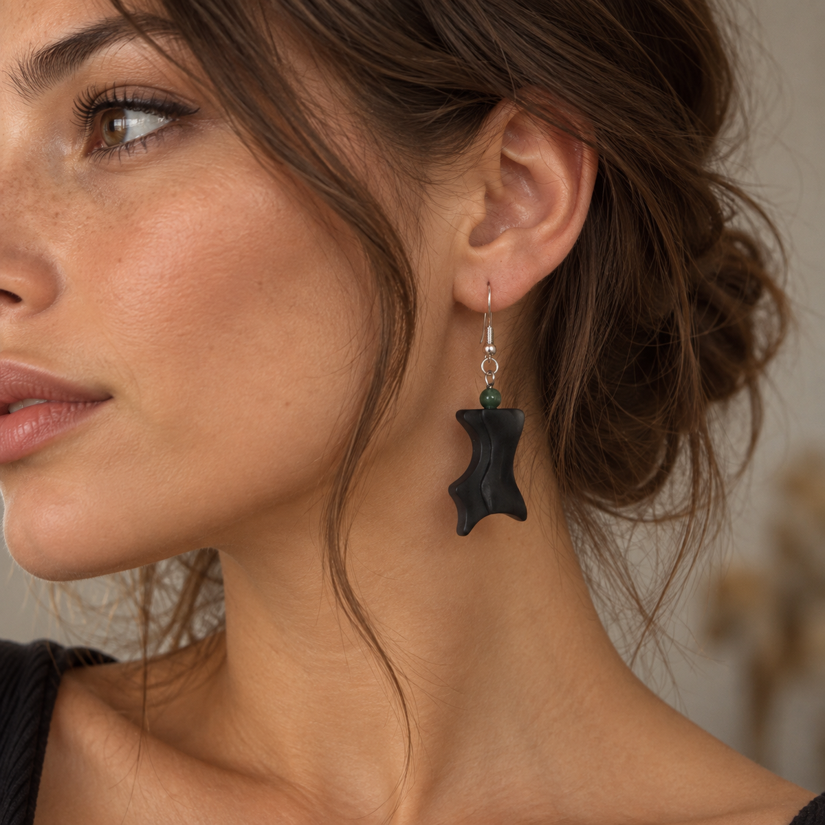 A close-up of a woman wearing a black abstract dangle earring with a small green bead and silver hook, shown against her ear and neck.