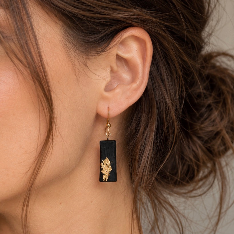 A close-up of a woman's ear wearing a dangling earring with a black rectangular wooden charm and a gold-toned floral design.