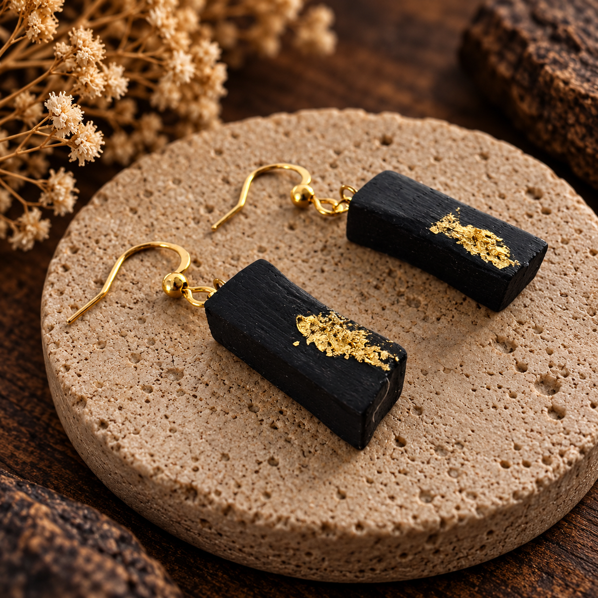 A pair of black rectangular drop earrings with textured gold leaf accents and gold fishhook wires, displayed on a round beige stone slab with dried flowers in the background.
