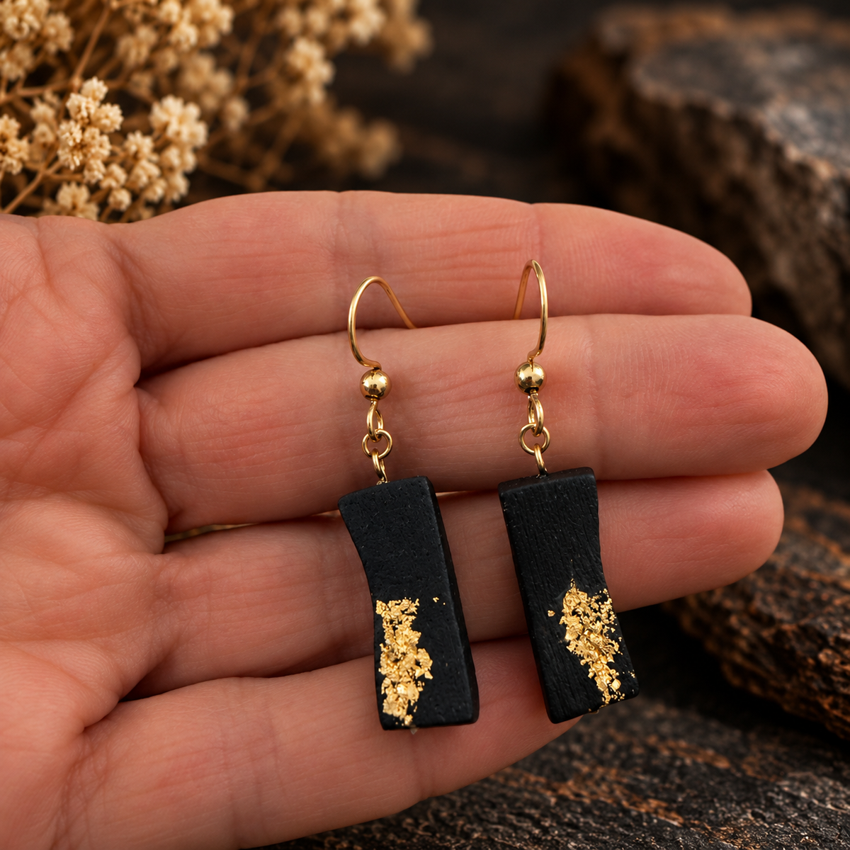 A pair of gold-tone drop earrings with rectangular black charms decorated with gold leaf, held in a person's hand against a rustic background.