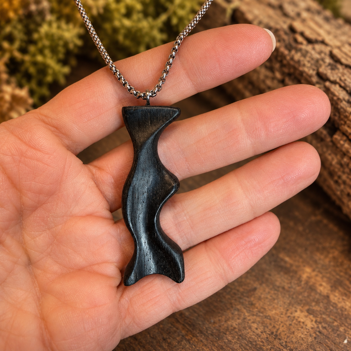 A hand holding a black wavy pendant necklace on a silver chain, with a rustic wood and moss background.