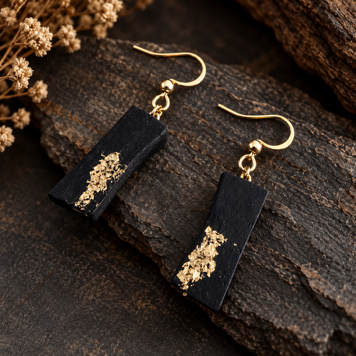 A pair of black rectangular drop earrings with gold fishhook wires and textured gold glitter accents, displayed on a dark rustic wood surface with dried flowers.