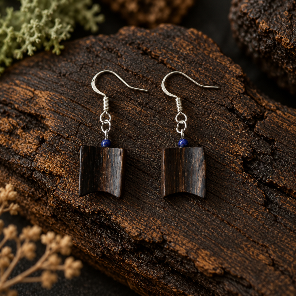 A pair of handmade wooden dangle earrings with dark brown wood pendants, silver fishhook wires, and small blue beads, displayed on a textured piece of dark wood.