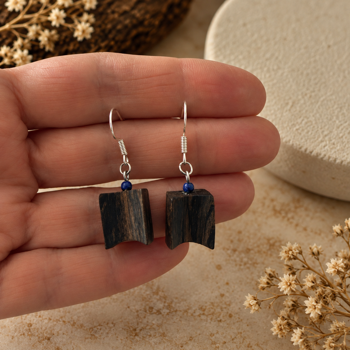 A pair of handmade dangle earrings with dark wood pendants and small blue beads, held on a person's fingers against a neutral background with dried flowers.