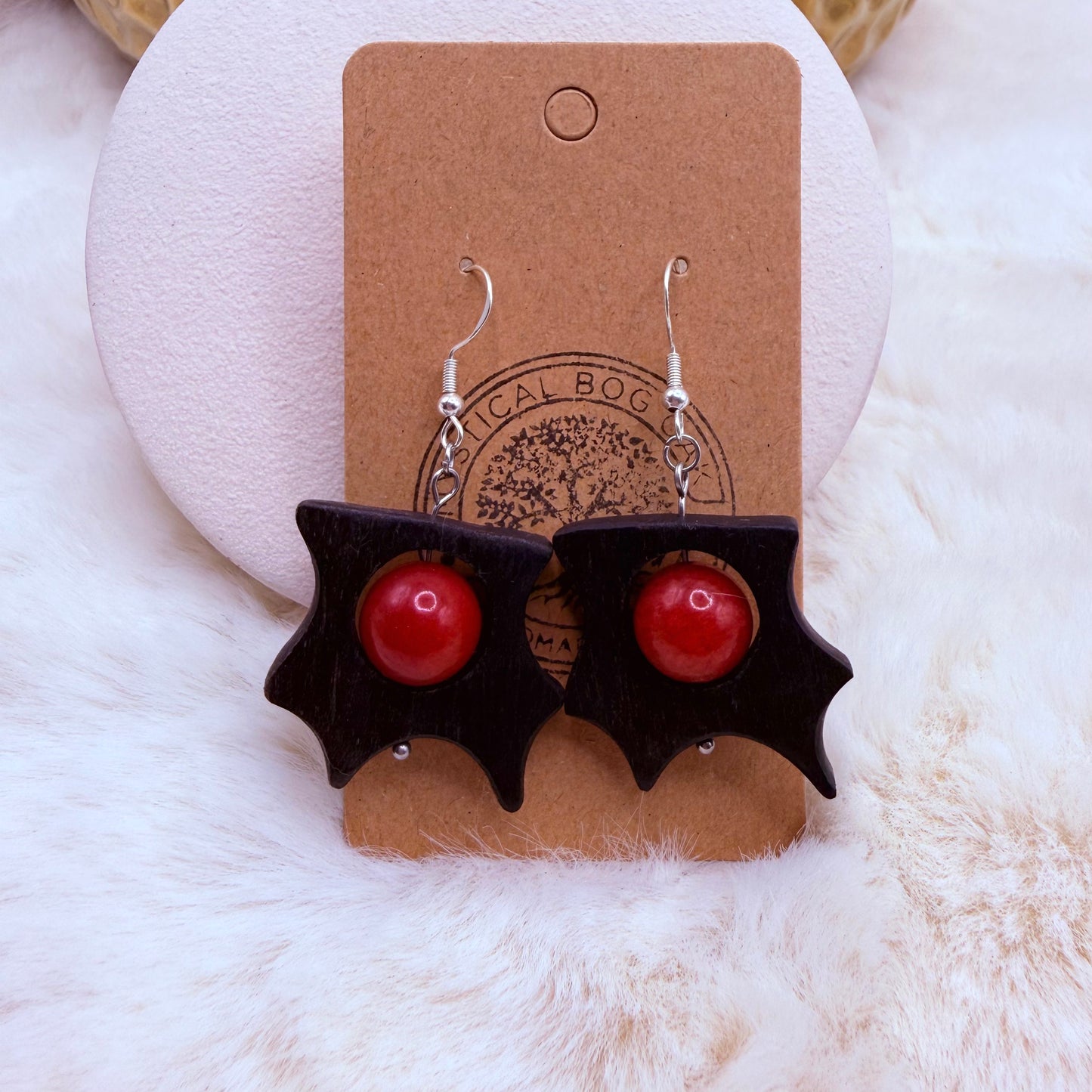 Pair of earrings with red balls and black bat wings on a cardboard backing with a brand logo.