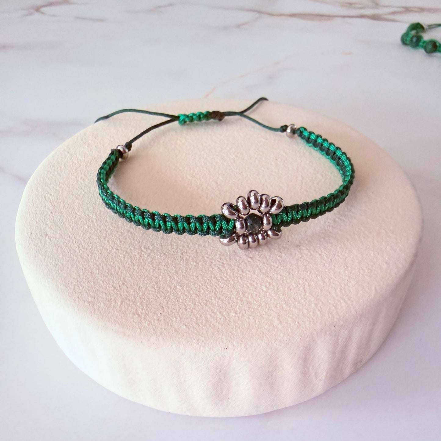 Green braided necklace with a silver flower pendant on a white cushion