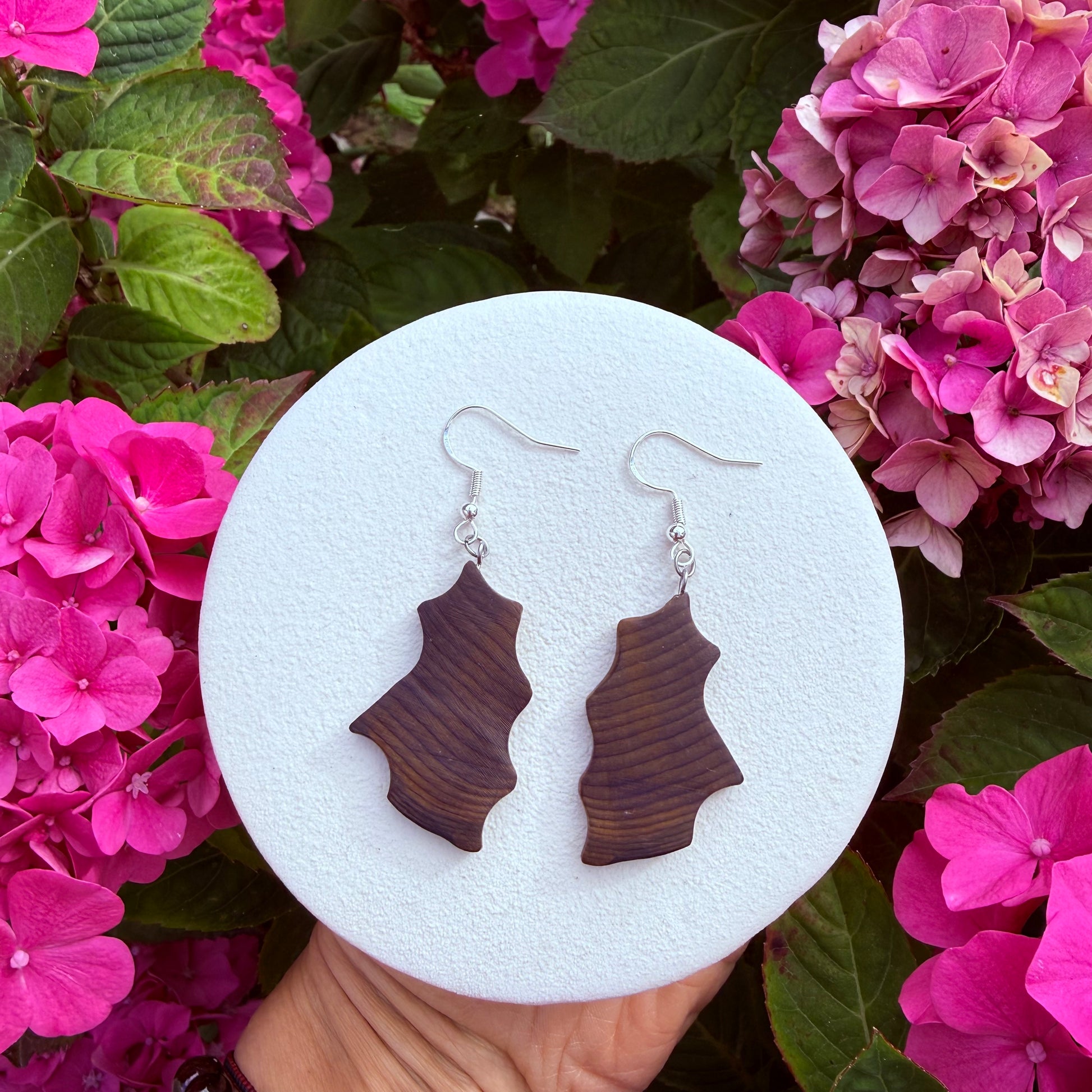 A pair of dark brown wavy wooden drop earrings with silver fishhook wires, displayed on a round white foam stand held by a hand against a background of pink hydrangea flowers.