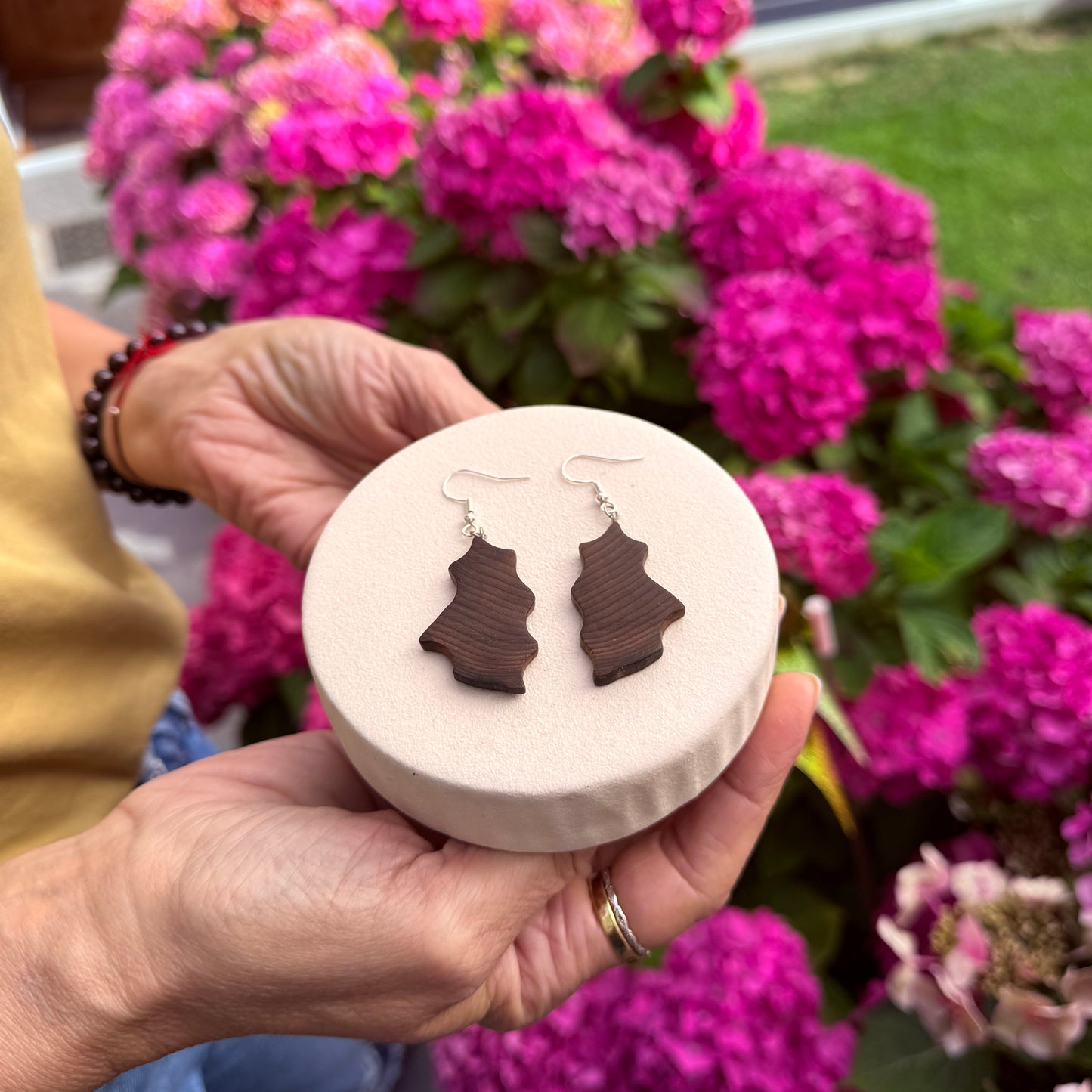 A person holds a round display stand showing a pair of dark brown wooden dangle earrings shaped like pine trees, with a blurred background of bright pink hydrangea flowers.