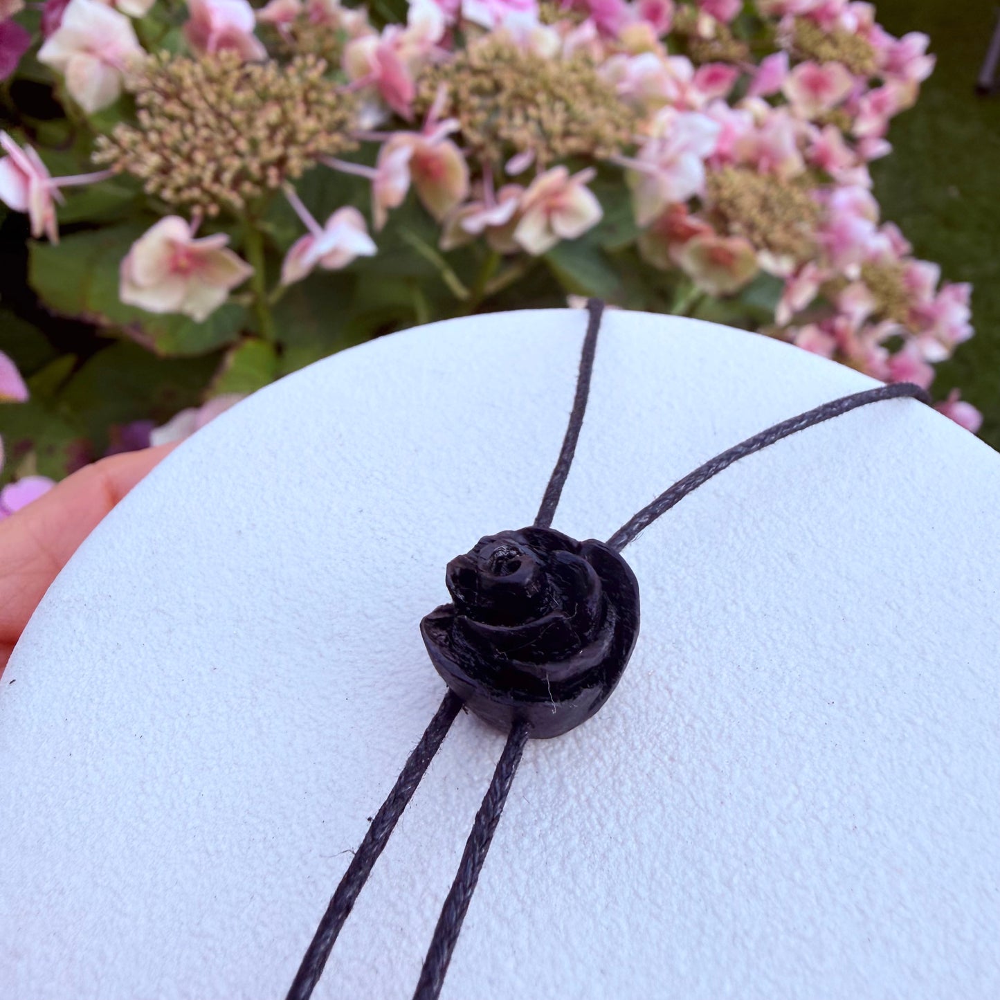 A black cord necklace with a sculpted black rose pendant, displayed on a white circular stand with a blurred background of pink flowers and green foliage.