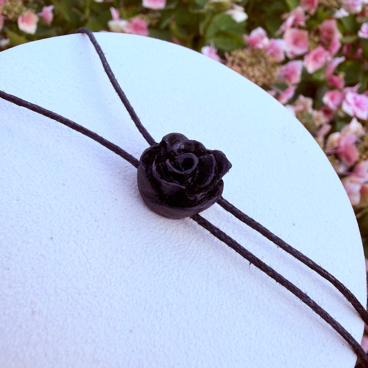 A black leather cord bracelet with a central black rose charm, displayed on a white circular stand with blurred pink flowers in the background.