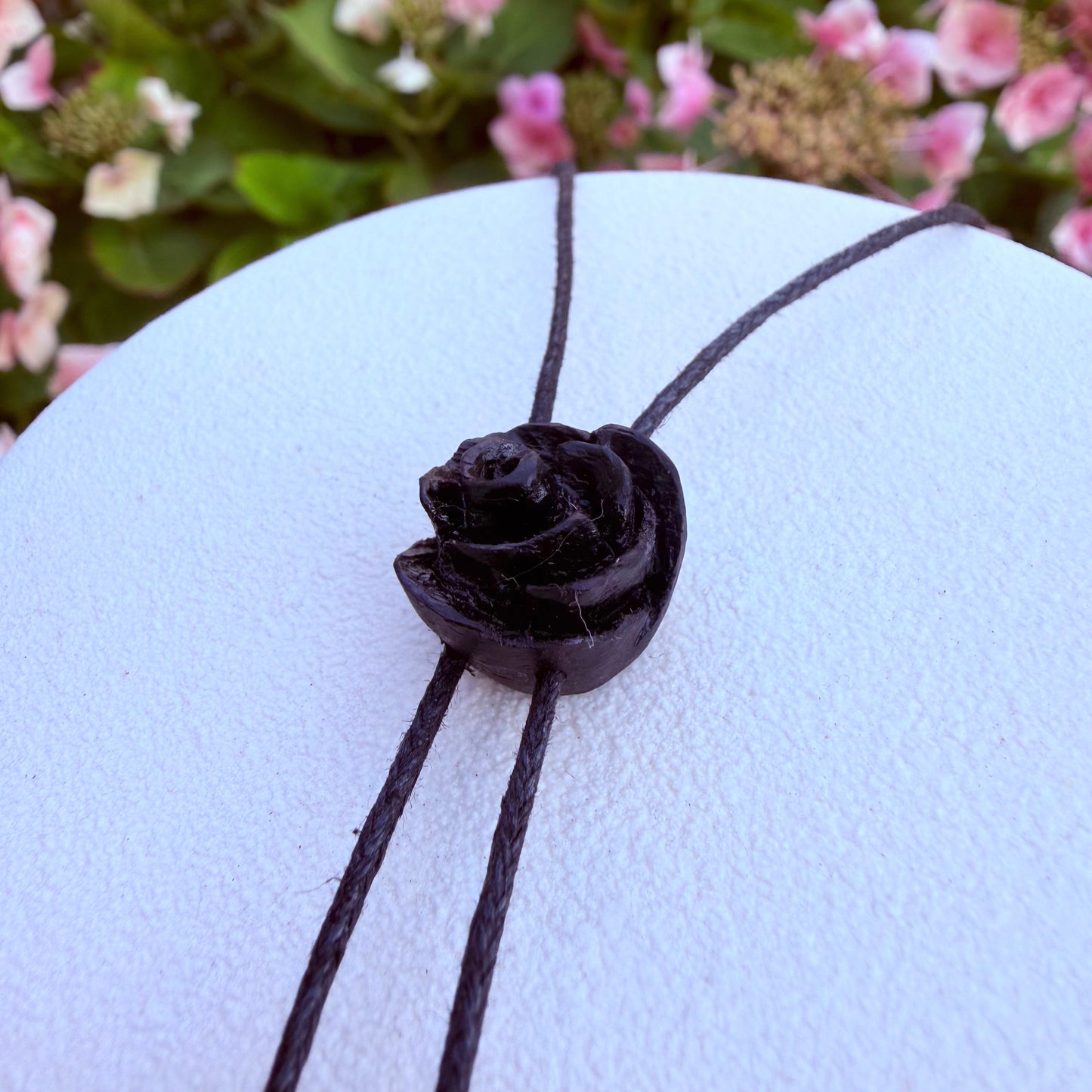 A close-up of a dark brown leather cord bolo tie with a carved rose-shaped bead, displayed on a white circular stand with blurred pink flowers in the background.