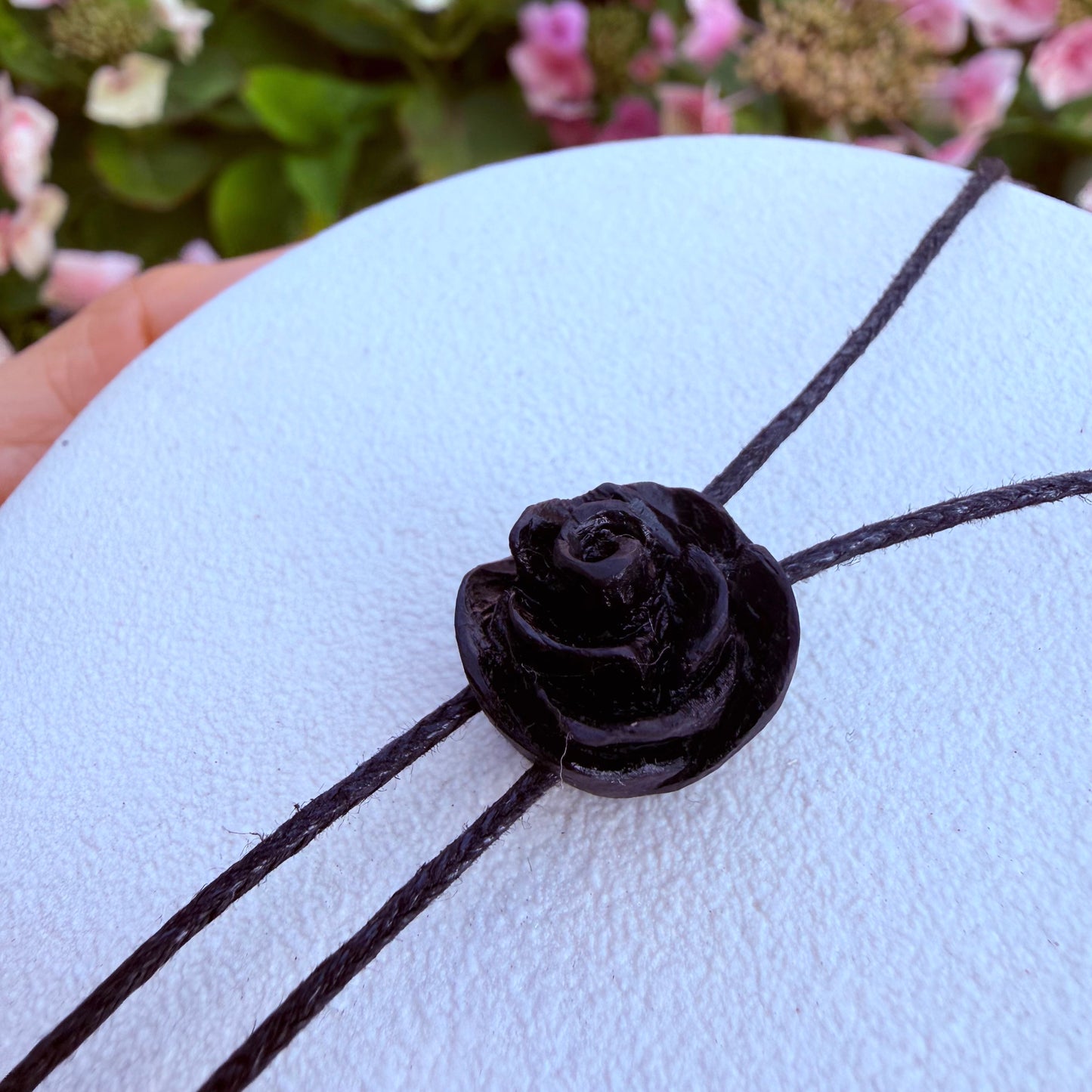 A close-up of a black cord necklace with a glossy black rose-shaped bead, displayed on a white textured surface with blurred pink flowers in the background.