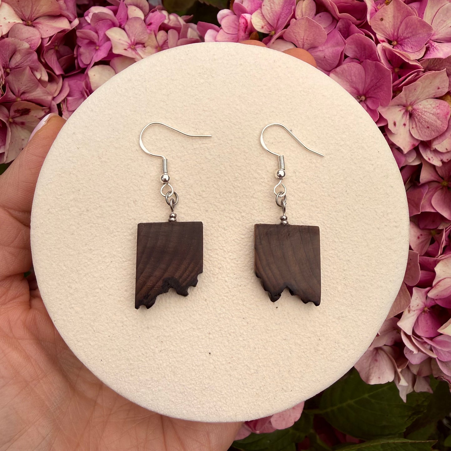 A pair of dangling earrings with dark brown wood cutouts shaped like state silhouettes, hanging from silver fishhook wires. The earrings are displayed on a round beige foam insert held by a hand in front of pink hydrangea flowers.