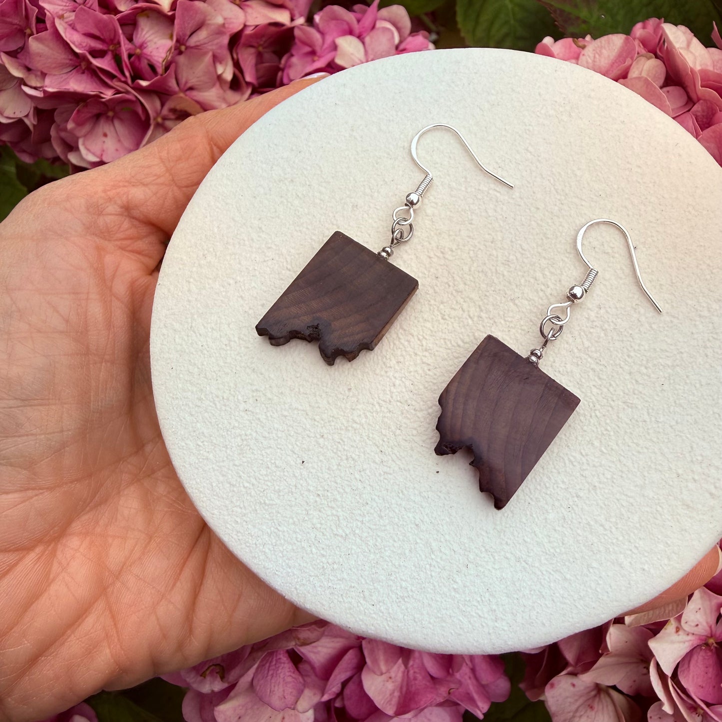 A pair of handmade drop earrings with dark brown wood inlays shaped like the state of Idaho, hanging from silver fishhook wires. The earrings are displayed on a round white foam stand held by a hand, with pink hydrangea flowers in the background.