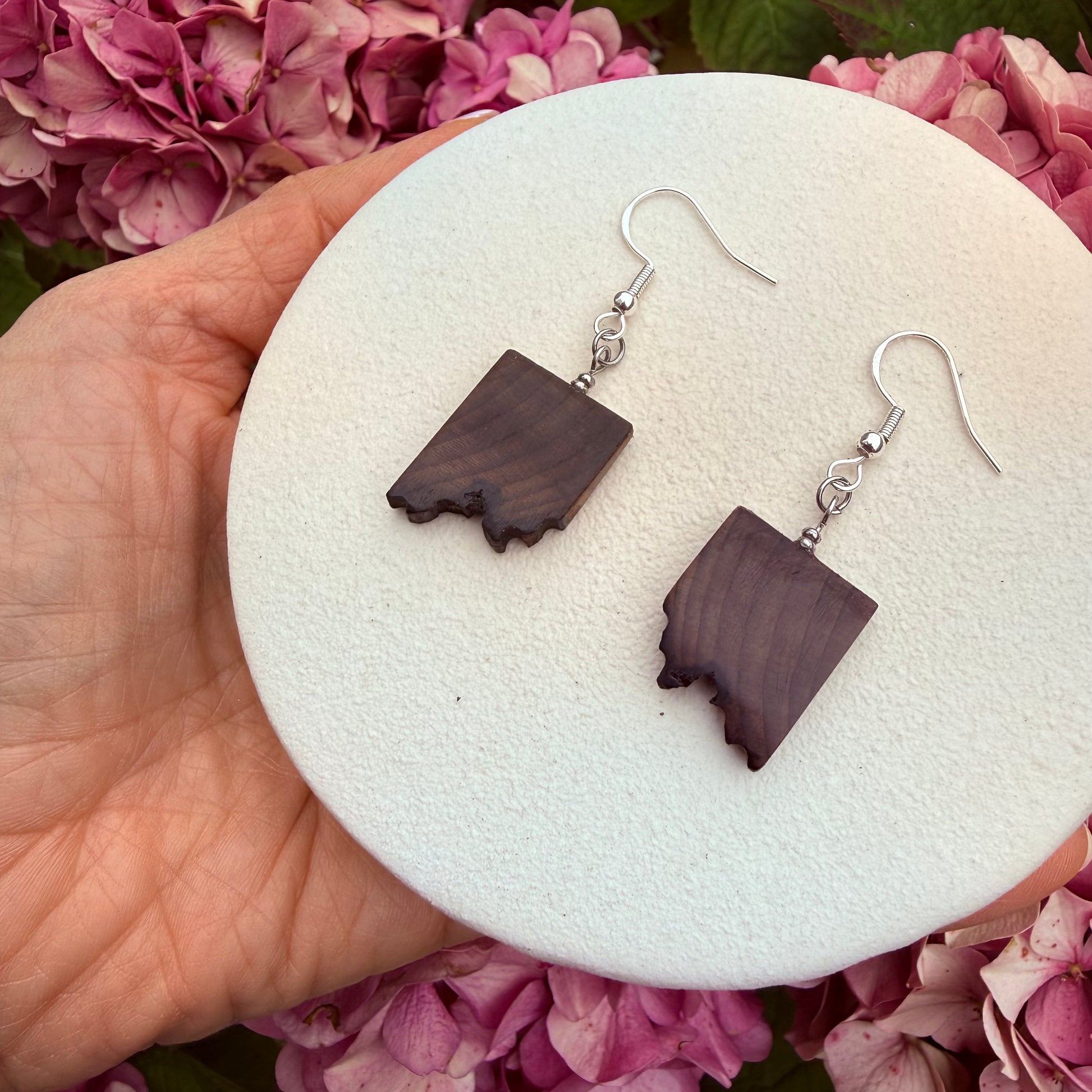 A pair of handmade drop earrings with dark brown wood inlays shaped like the state of Idaho, hanging from silver fishhook wires. The earrings are displayed on a round white foam stand held by a hand, with pink hydrangea flowers in the background.