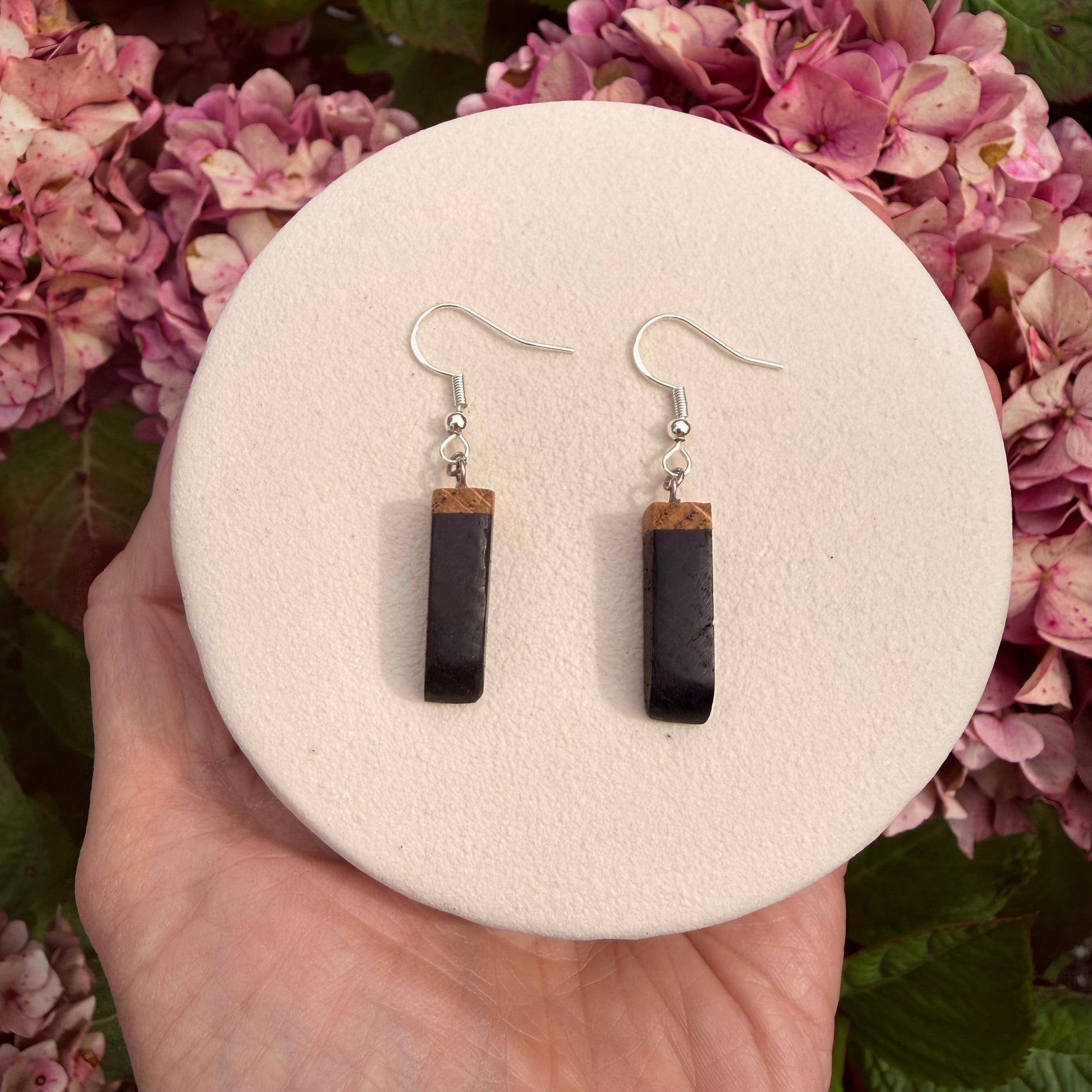 A pair of drop earrings with dark rectangular stone pendants and small natural wood tops, hanging from silver fishhook wires. They are displayed on a round cream-colored stand held in a hand, with pink hydrangeas in the background.