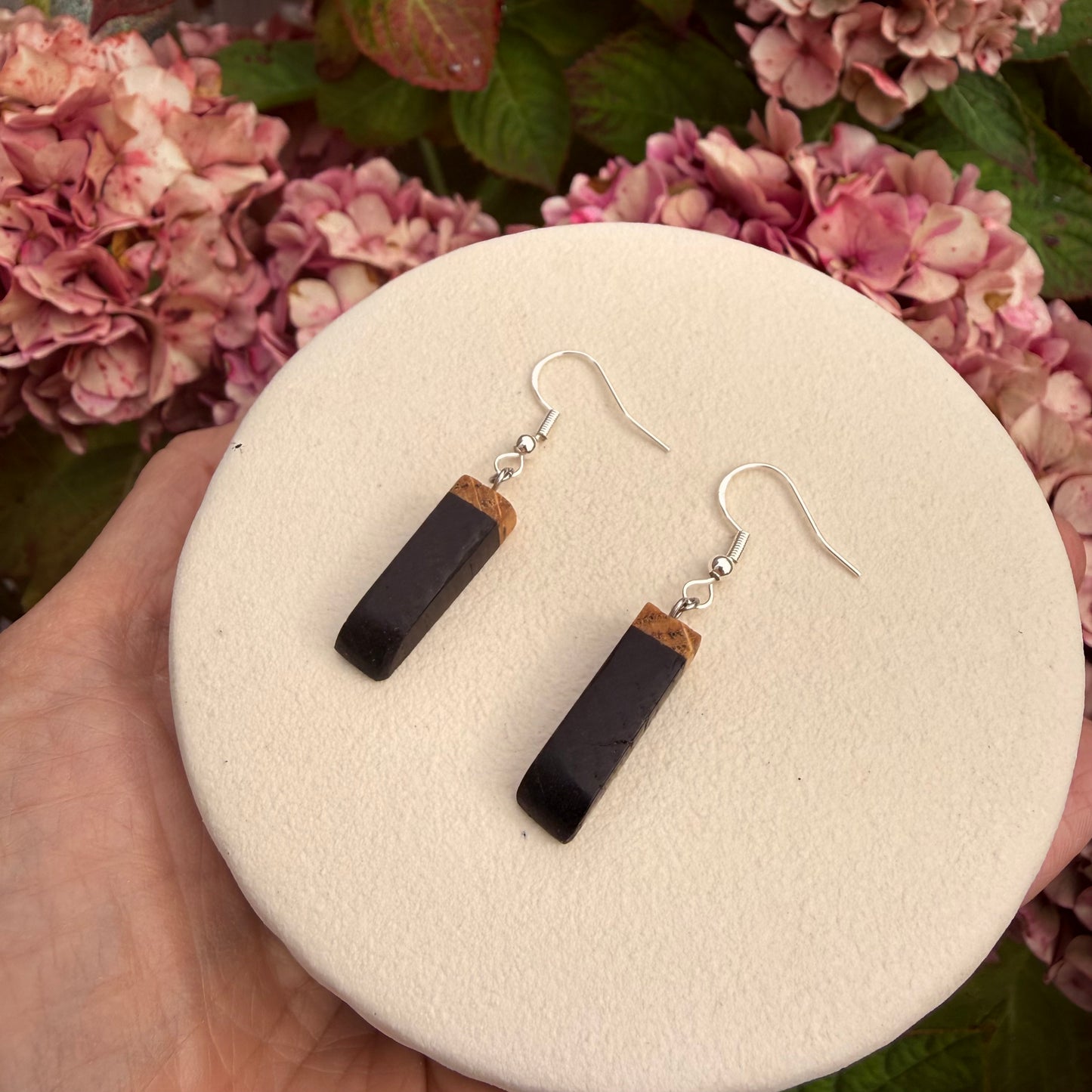 A pair of drop earrings with rectangular black stone pendants and small tan wood sections, hanging from silver fishhook wires, displayed on a round cream foam pad held in a hand with pink flowers in the background.