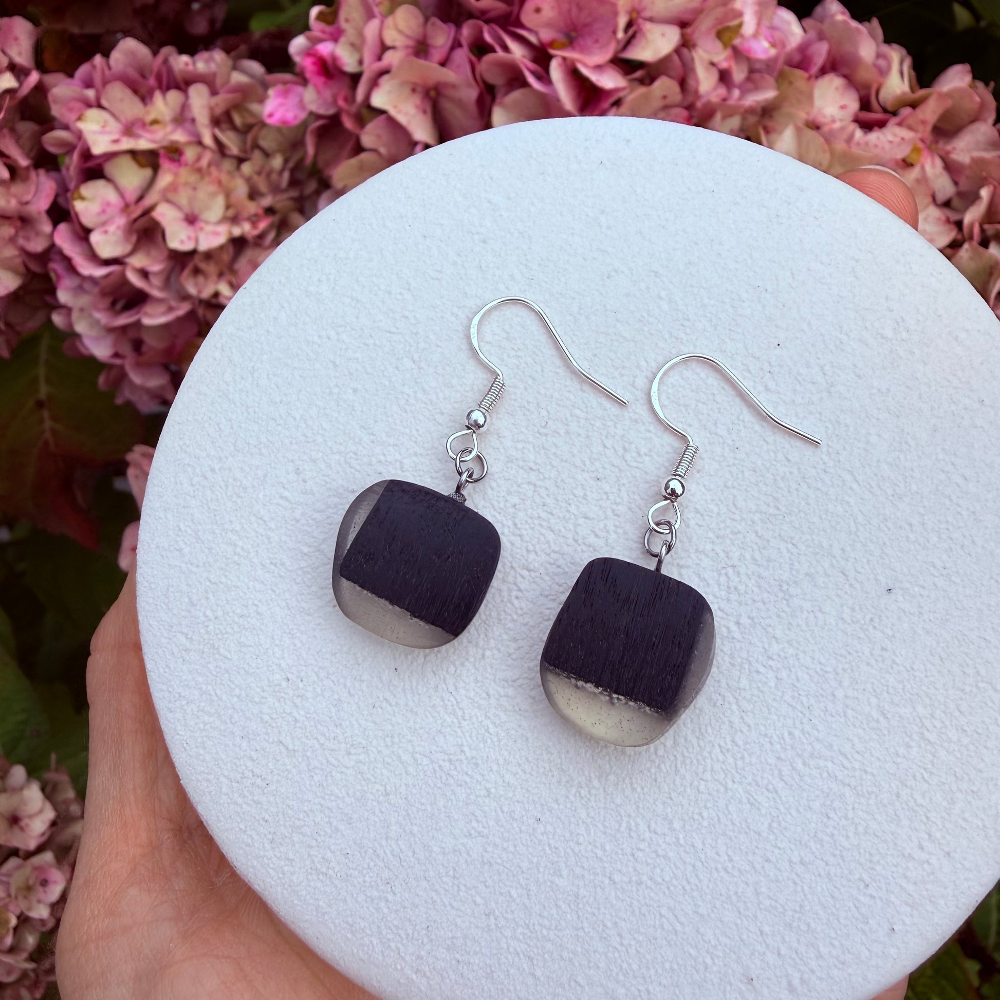 A pair of handmade dangle earrings with square dark brown wooden pendants and silver fishhook wires, held on a white foam display by a hand against a background of pink hydrangeas.