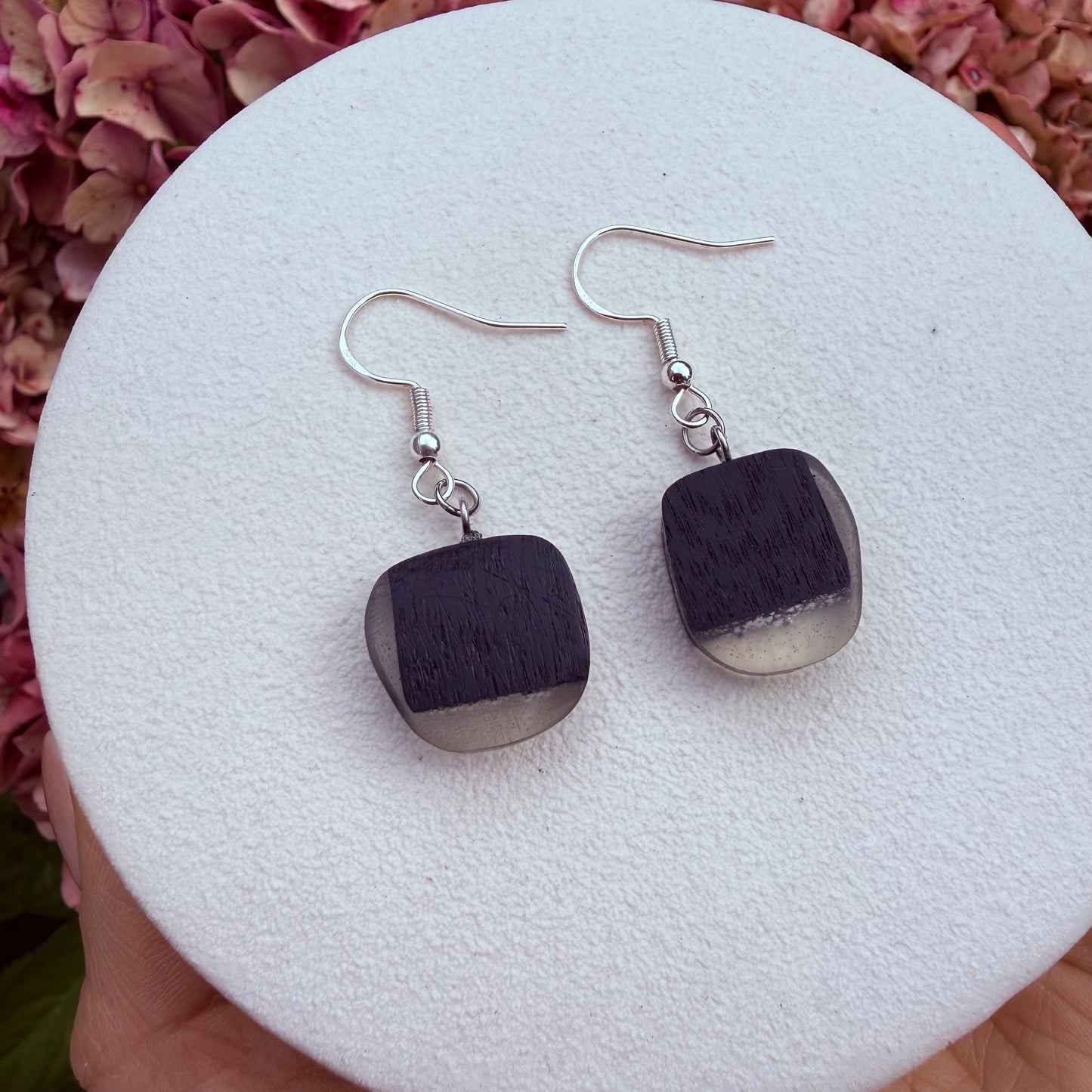 A pair of handmade dangle earrings with square dark gray wooden pendants and silver fishhook wires, resting on a white textured display pad held by a hand.