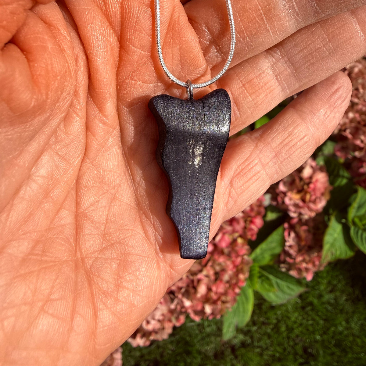 A dark, polished stone pendant with a natural organic shape hanging from a silver chain, held in a person's hand with pink flowers and green foliage in the background.