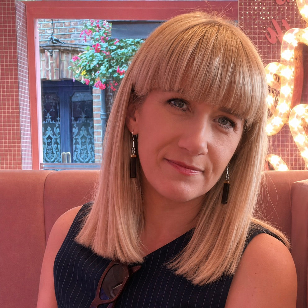 A woman with blonde shoulder-length hair and straight bangs, wearing a dark sleeveless top and dangling earrings, seated in a pink booth in a restaurant or cafe setting.