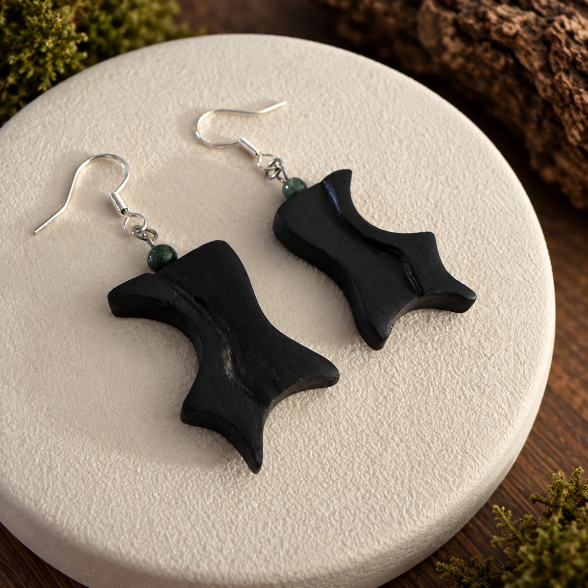 A pair of black dangle earrings with silver fishhook wires and small dark green beads, displayed on a round textured white plate over a wooden surface.