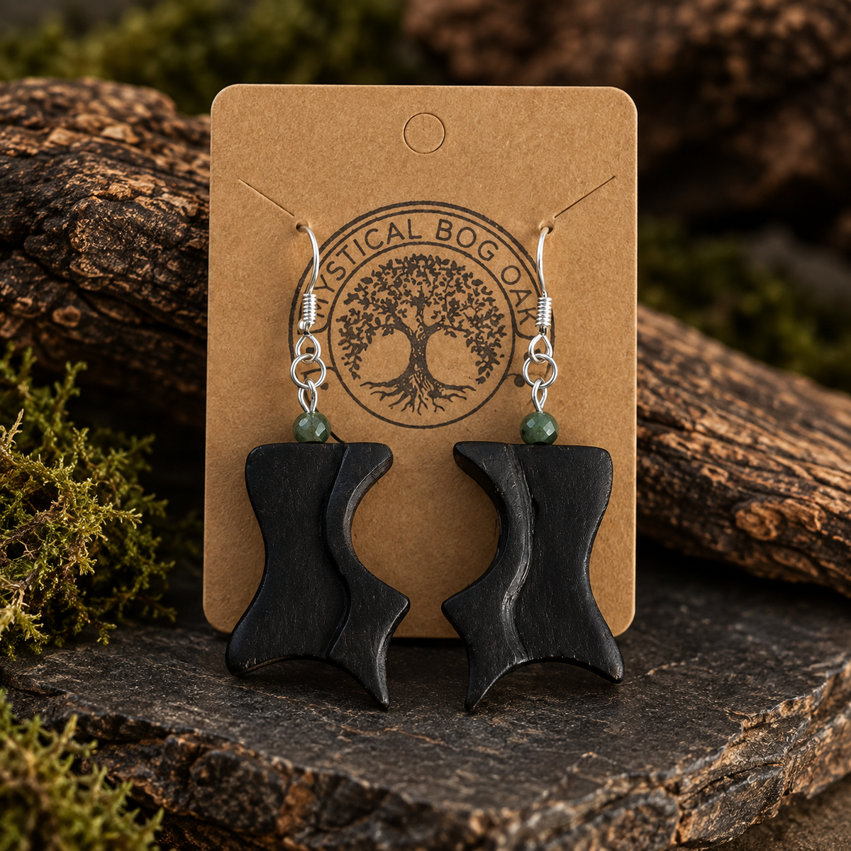 A pair of black wooden dangle earrings with small green bead accents and silver fishhook wires, displayed on a brown kraft card printed with a tree logo. The card is set against a rustic background of moss, logs, and dark wood.