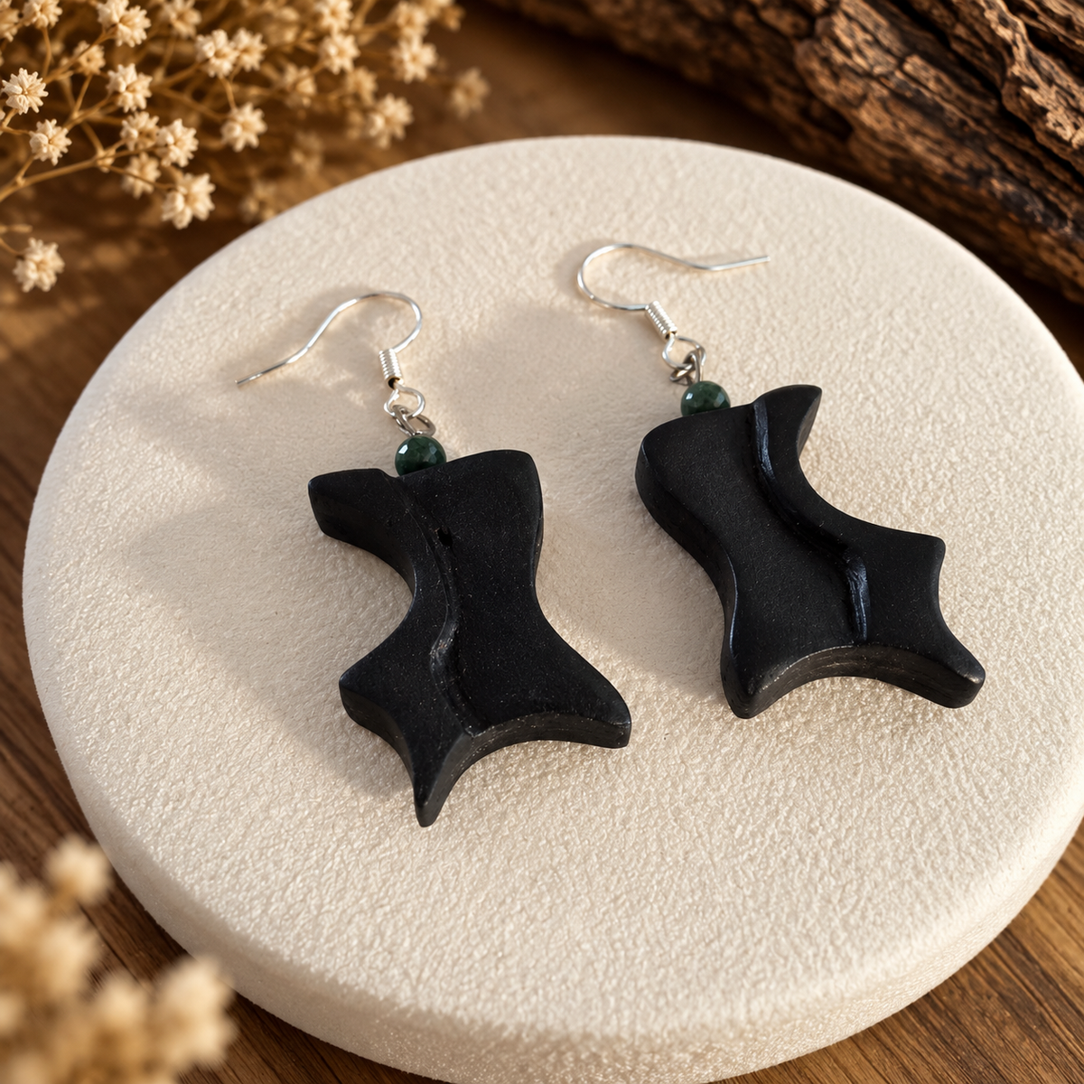 A pair of black abstract sculptural dangle earrings with silver fishhook wires and small dark green beads, displayed on a round textured cream plate over a wooden surface with dried flowers.