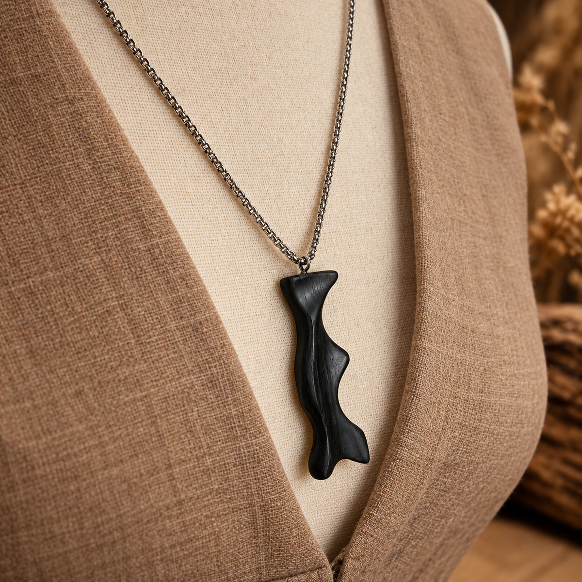A black organic-shaped pendant necklace on a silver chain, displayed on a beige linen dress form with a brown textured background.