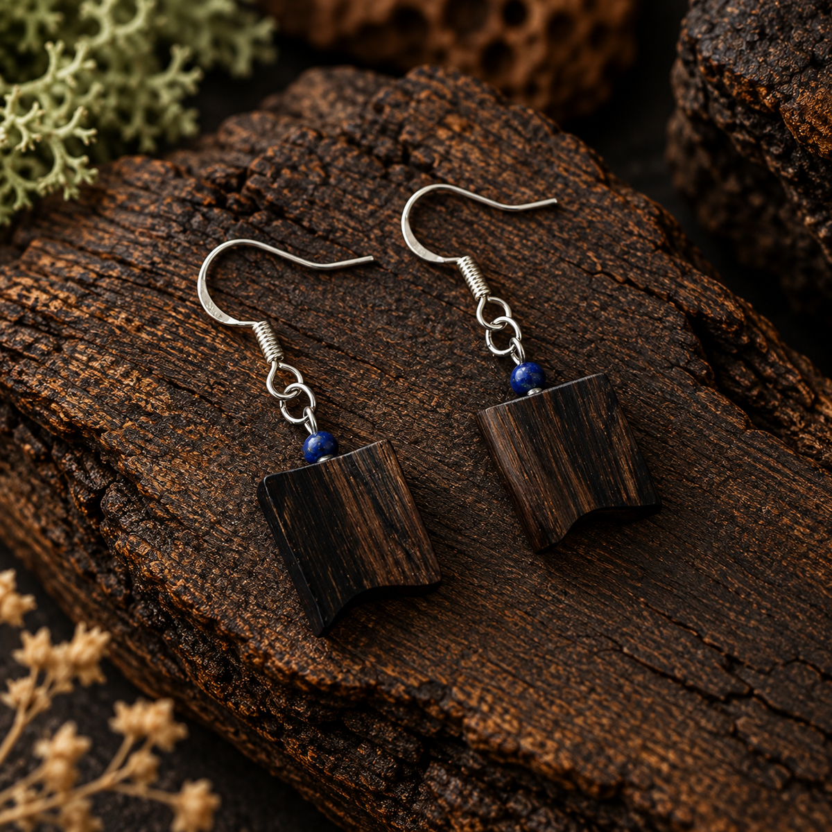 A pair of handmade drop earrings featuring dark brown wood grain rectangular pendants with small blue beads and silver fishhook wires, displayed on a textured dark wood log.