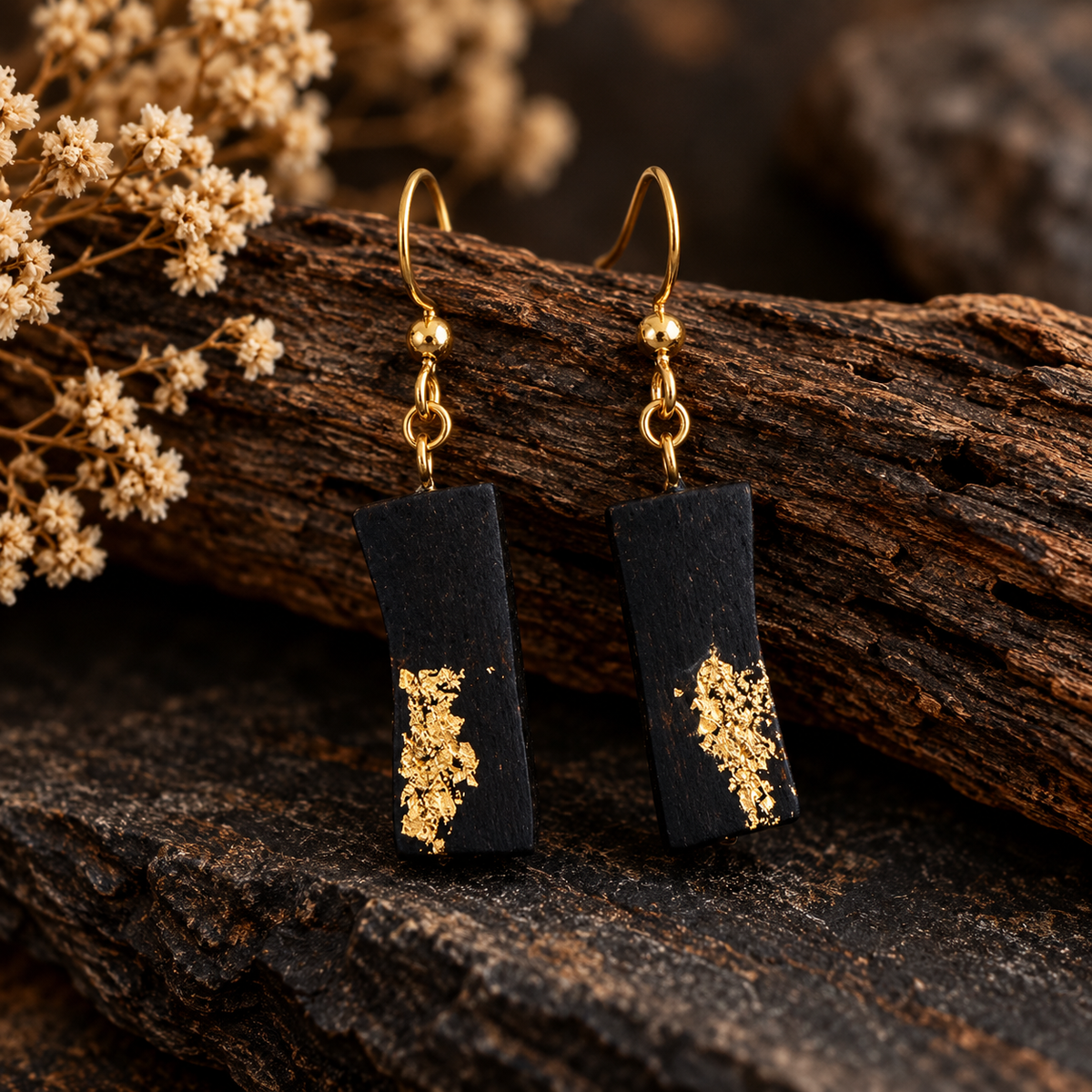 A pair of rectangular black drop earrings with gold leaf accents and gold fishhook wires, displayed on a piece of dark wood with dried flowers.