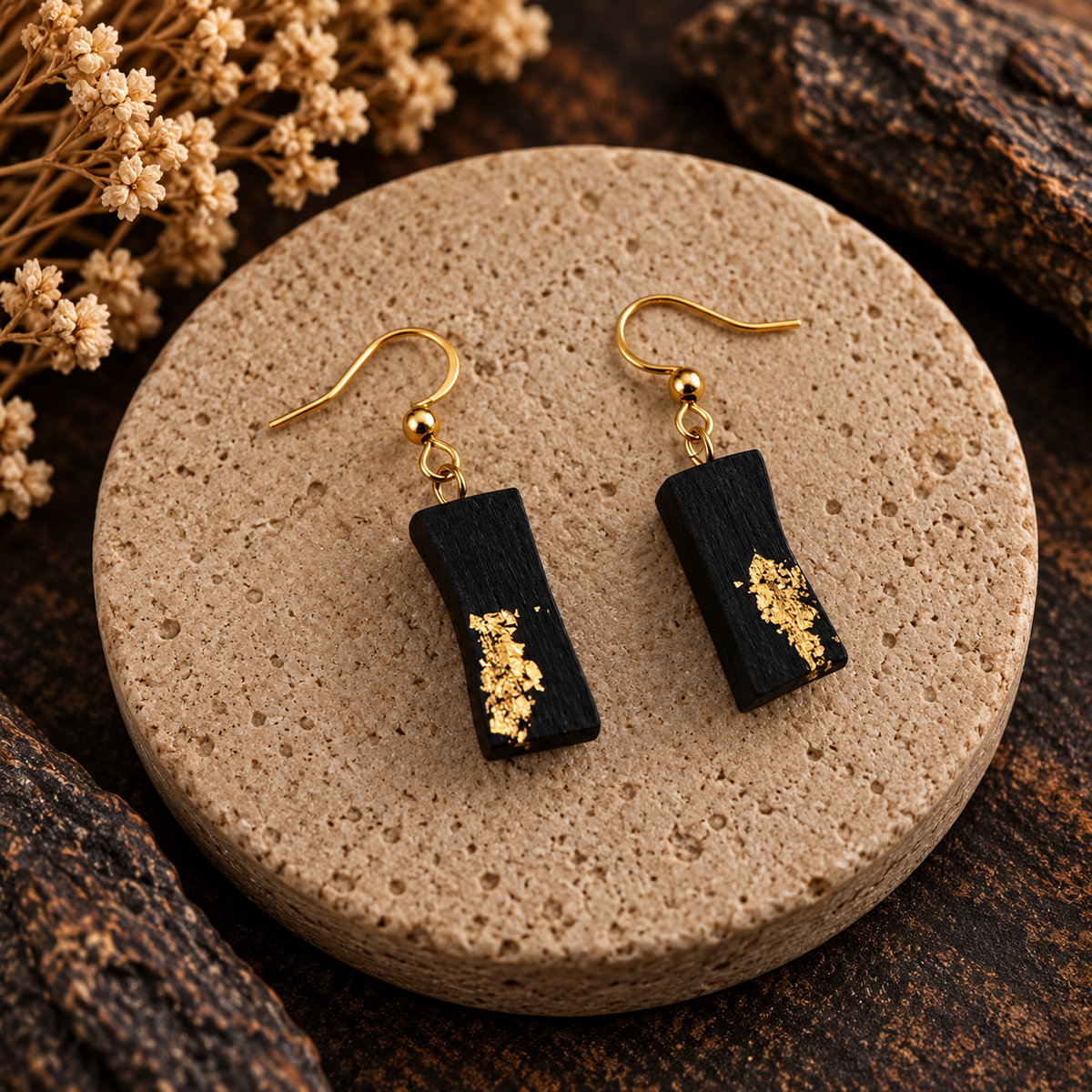 A pair of black rectangular drop earrings with gold fishhook wires and gold leaf accents, displayed on a round beige stone plate with dried flowers and dark wood in the background.