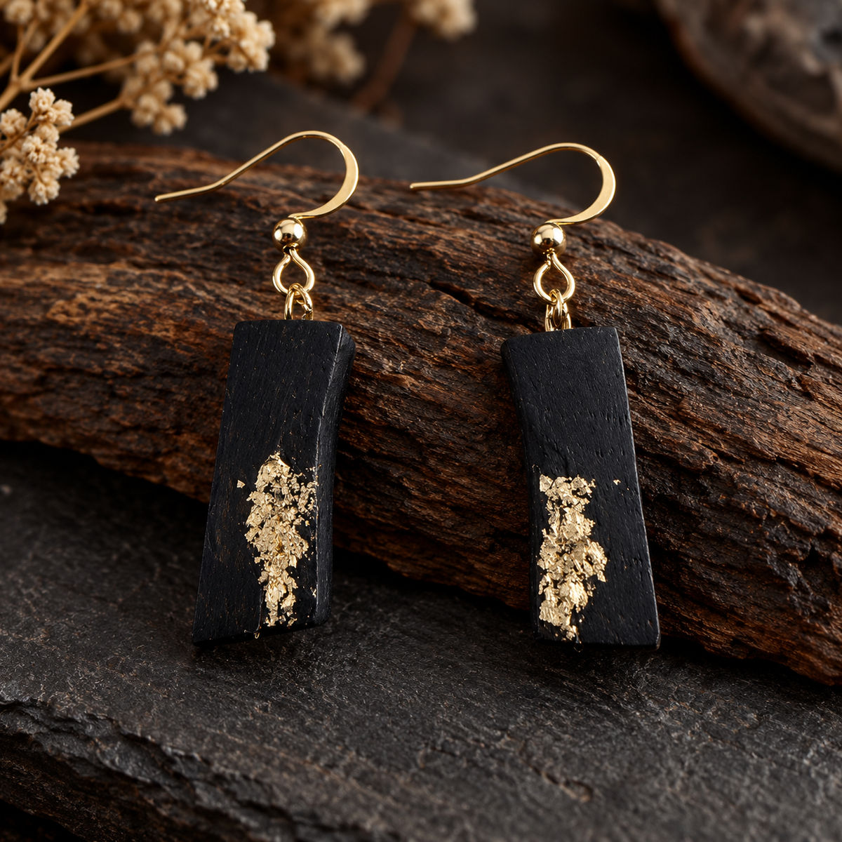A pair of rectangular black drop earrings with textured gold leaf inlay and gold fishhook wires, displayed on a dark rustic wood surface.