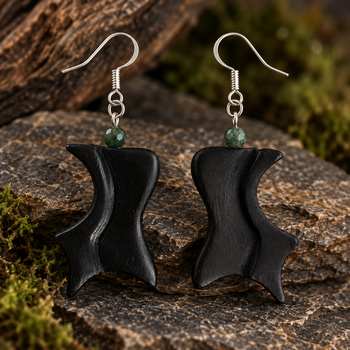A pair of black wooden dangle earrings with silver fishhook wires and small green faceted beads, displayed on a textured rock with moss and wood in the background.