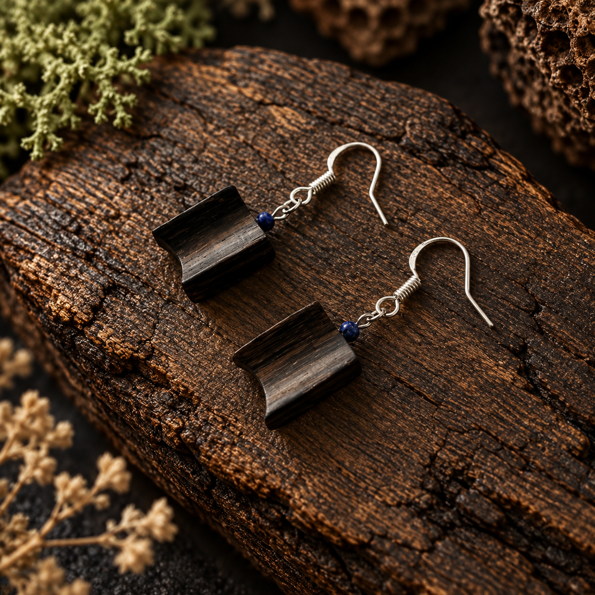 A pair of handmade dangle earrings with dark brown wood slice pendants and small blue bead accents, displayed on a textured dark wood log with dried botanicals.