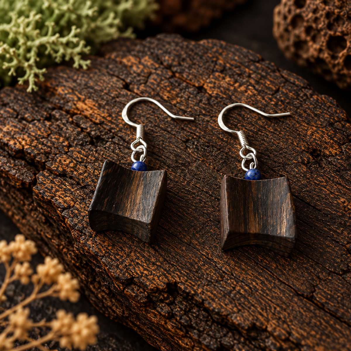A pair of handmade geometric drop earrings featuring dark brown wood cubes with visible grain, suspended from silver fishhook wires with small blue bead accents, displayed on a textured dark wood log.