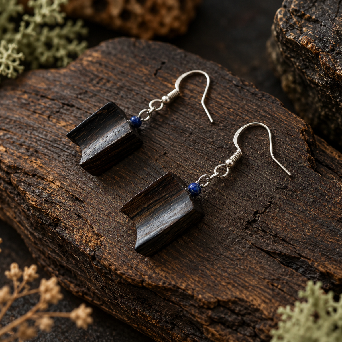 A pair of handmade dangle earrings with dark wood blocks and small blue beads, displayed on a rustic piece of wood.