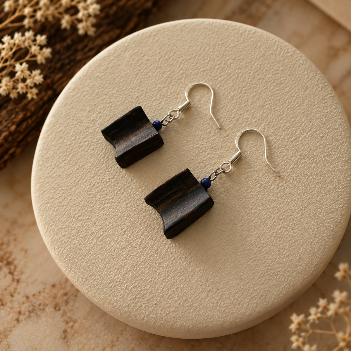 A pair of handmade dangle earrings with dark brown curved wooden pendants and small blue beads, hanging from silver fishhook wires on a textured beige round display plate.